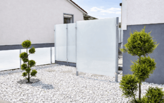 Moderner minimalistischer Vorgarten mit weißen Zierkieseln, kunstvoll geschnittenen Formschnittbäumen und satinierten Glas-Sichtschutzwänden zwischen modernen Häusern
