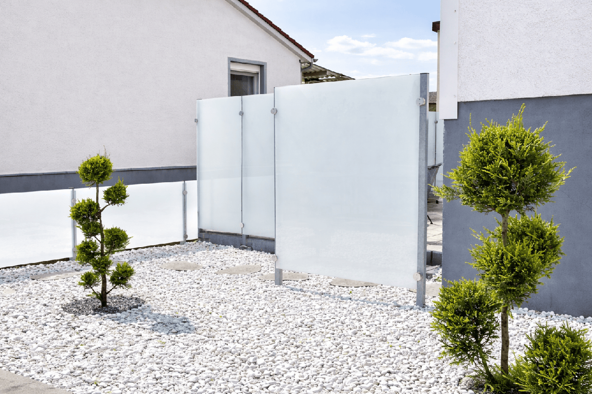 Moderner minimalistischer Vorgarten mit weißen Zierkieseln, kunstvoll geschnittenen Formschnittbäumen und satinierten Glas-Sichtschutzwänden zwischen modernen Häusern