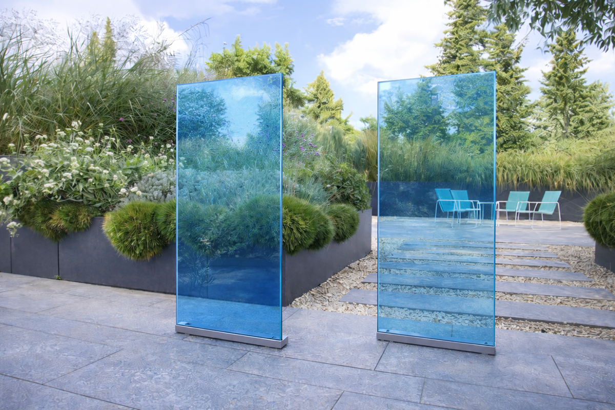 Moderner Garten mit blauen Glas-Sichtschutzpaneelen, üppiger Bepflanzung, Ziergräsern und einer stilvollen Terrasse mit Sitzmöbeln im Hintergrund.